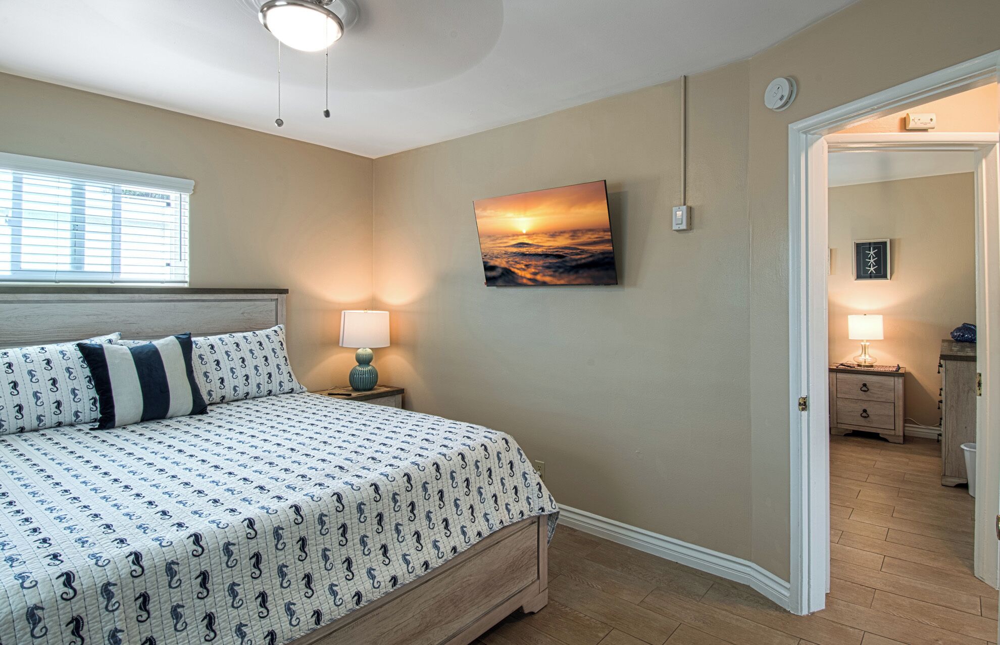 A bedroom with a bed, a nightstand, a wall-mounted lamp, a painting of a sunset, and a door leading to another room.