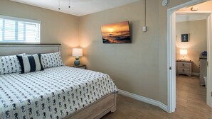 Un dormitorio con una cama, una mesita de noche, una lámpara de pared, un cuadro de un atardecer y una puerta que da a otra habitación.