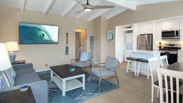 Una sala de estar moderna con un televisor grande, un comedor y una cocina con armarios blancos y electrodomésticos de acero inoxidable.