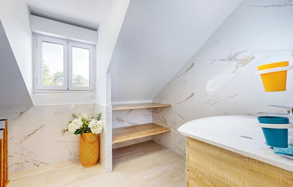A modern bathroom with a skylight, marble tiles, a wooden shelf, a white sink, and a yellow vase with flowers.