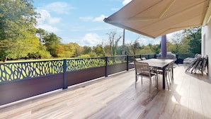 Un patio techado con mesa y sillas, una parrilla y vistas a árboles y una valla.