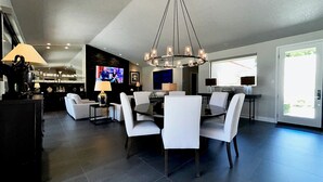 Een moderne eetruimte met een ronde tafel en witte stoelen, een kroonluchter en een televisie op de achtergrond.