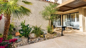 A patio with a stone floor, surrounded by various plants and a cactus, with outdoor furniture and a pergola. - Racquet Club Retreat | Modern Luxe Living in a Peaceful Desert Escape – managed by Greenday (Rancho Mirage)