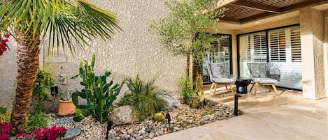 A patio with a stone floor, surrounded by various plants and a cactus, with outdoor furniture and a pergola.