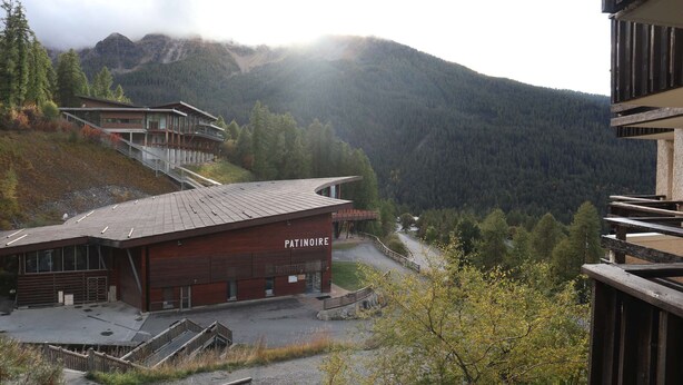 Un edificio moderno chiamato 'Patinoire', con tetto spiovente, circondato da un paesaggio montuoso e una fitta foresta.