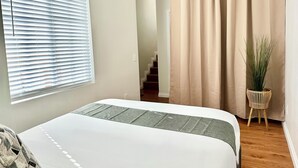 A bedroom with a bed, a window with blinds, a curtain, and a plant on a wooden stool. - Disneyland Getaway Tri-level Gated Townhome (Placentia)