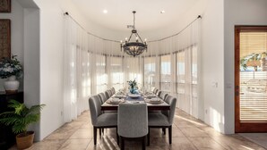 A dining area with a long table set for six, surrounded by sheer curtains, a chandelier, and potted plants. - The Grand Oasis – Resort-Style Retreat w/ Sauna, Cold Plunge, Mini Golf & Volleyball (Goodyear)