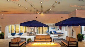 A spacious indoor lounge area with a central fire feature, multiple seating options including sofas and chairs, and overhead string lights. - The Grand Oasis – Resort-Style Retreat w/ Sauna, Cold Plunge, Mini Golf & Volleyball (Goodyear)
