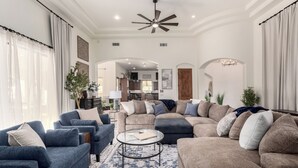 A spacious living room with a large sectional sofa, a smaller sofa, and a coffee table. There is a ceiling fan and a chandelier. - The Grand Oasis – Resort-Style Retreat w/ Sauna, Cold Plunge, Mini Golf & Volleyball (Goodyear)