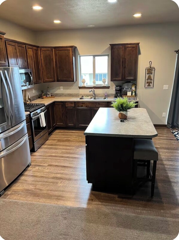 Cocina con armarios de madera, electrodomésticos de acero inoxidable, una isla central con asientos y un ventanal que proporciona luz natural.