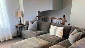 A large sectional sofa with multiple cushions, a small side table with a lamp, and a staircase in the background.