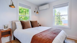 Un dormitorio con una cama, dos lámparas de noche, una mesita, un cuadro enmarcado y una ventana con vista a árboles y al mar.