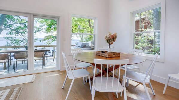 A dining area with a round wooden table, white chairs, and a view of the outdoors through large windows.
