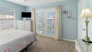 A bedroom with a bed, a television, a ceiling fan, a window with blinds, a door with curtains, and a small table with a lamp. - "Mermaid Cove" offers canal views, fenced-in yard, boat parking, pet friendly! (Mexico Beach)