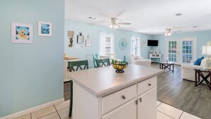 A spacious living area with a kitchen island, dining table, and chairs. There are ceiling fans, a TV, and artwork on the walls. - "Mermaid Cove" offers canal views, fenced-in yard, boat parking, pet friendly! (Mexico Beach)