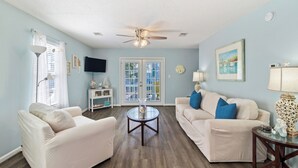 A living room with two white sofas, a ceiling fan, and a large window with blinds. - "Mermaid Cove" offers canal views, fenced-in yard, boat parking, pet friendly! (Mexico Beach)