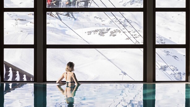 Eine Person im Bikini steht an einem Fenster mit Blick auf eine verschneite Landschaft, wobei ein Pool die Szenerie reflektiert.