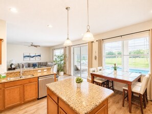 A modern kitchen with granite countertops, a dining area with a glass table, and a view of a pool through large windows.