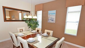 Une salle à manger avec une table rectangulaire dressée pour quatre personnes, entourée de chaises blanches. Un grand miroir est accroché au-dessus de la table, et il y a deux fenêtres équipées de stores.