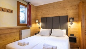 Cama doble con sábanas y toallas blancas, paredes de madera y dos lámparas de noche, además de una ventana con cortina.