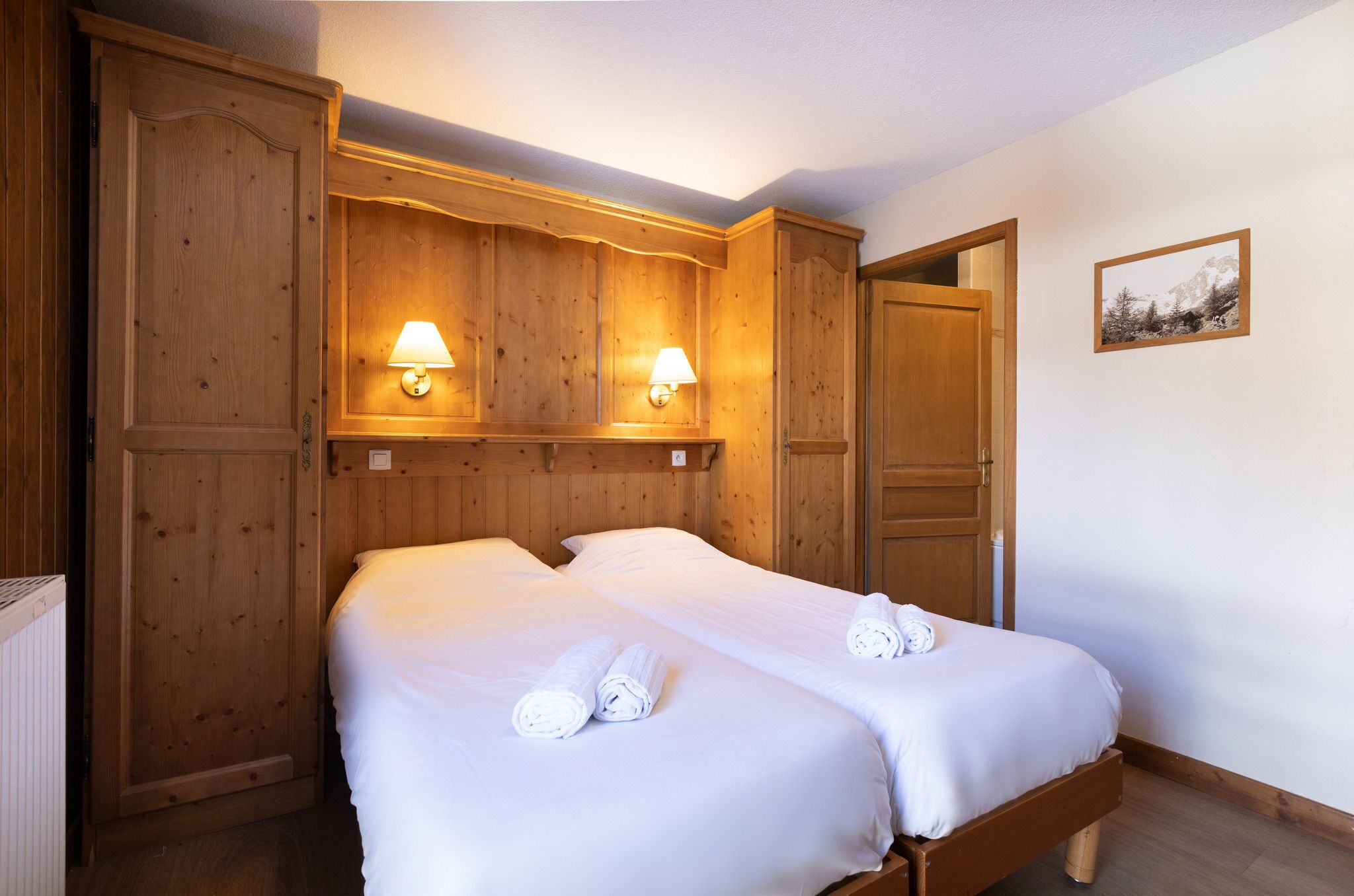 A double bed with white linens and rolled towels, wooden paneling on the walls, two wall-mounted lamps, and a framed picture on the wall.