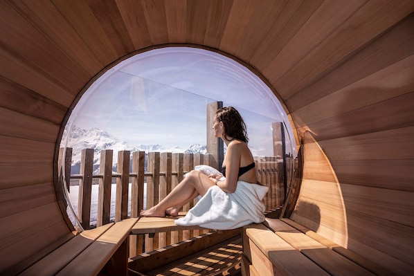 Un sauna en bois dot é d’une fenêtre circulaire, de bancs en bois et offrant une vue sur des montagnes enneigées.