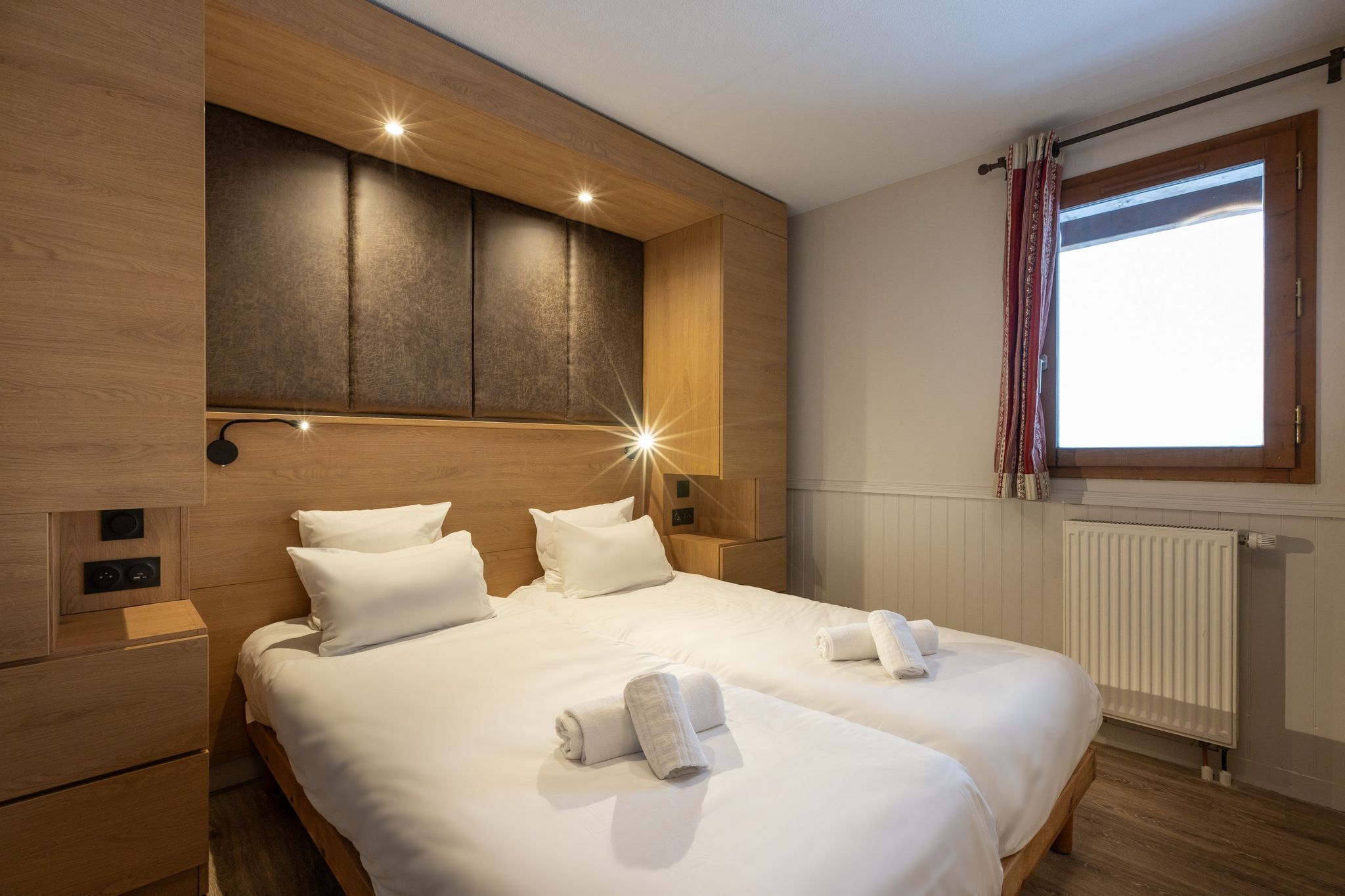 A double bed with white linens, a wooden headboard, bedside tables, and a window with curtains.