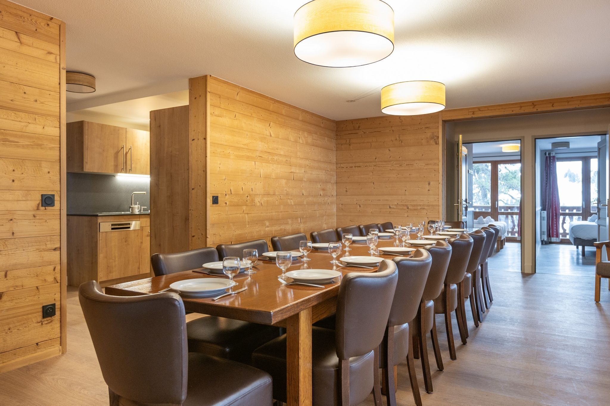 A long wooden dining table with chairs, a kitchen area, and a view of the outdoors.
