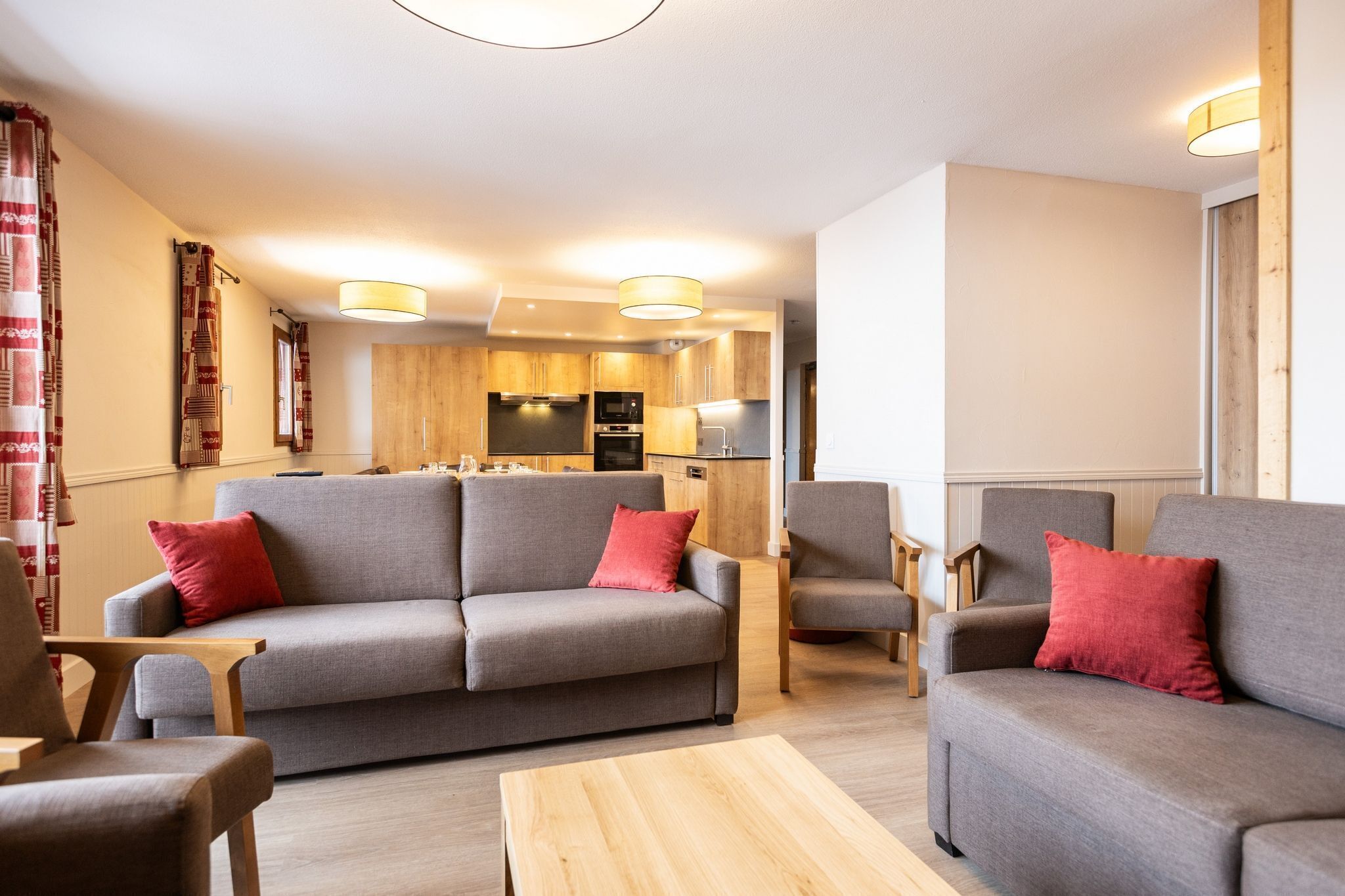 A modern living room with a grey sofa and armchairs, wooden flooring, and a kitchen in the background.