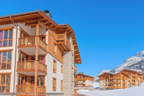 Un edificio de madera de varios pisos con balcones, pilares de piedra y una montaña nevada al fondo.