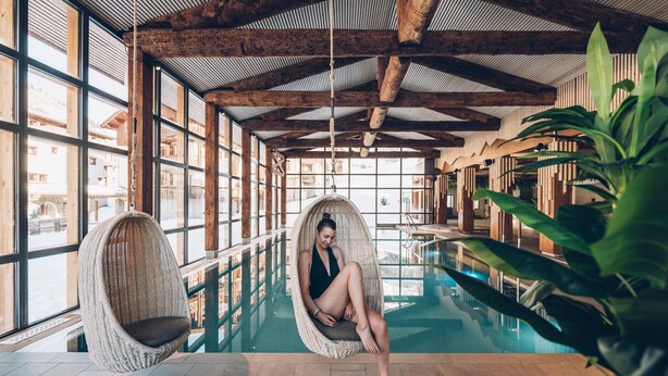 Ein großes Hallenbad mit Holzbalkendecke und gro ßen Fenstern. Zwei Korbsessel stehen am Pool.