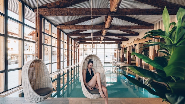 Une grande piscine intérieure avec un plafond en bois et de grandes fenêtres. Deux fauteuils en osier sont placés au bord de la piscine.