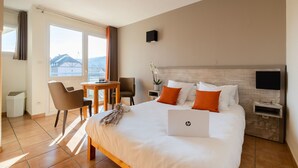A hotel room with a large bed, a desk, and a chair. There is a laptop on the bed, and a TV on the wall.