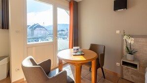 A modern room with a round wooden table, two chairs, a potted orchid, and a view of buildings through a large window.