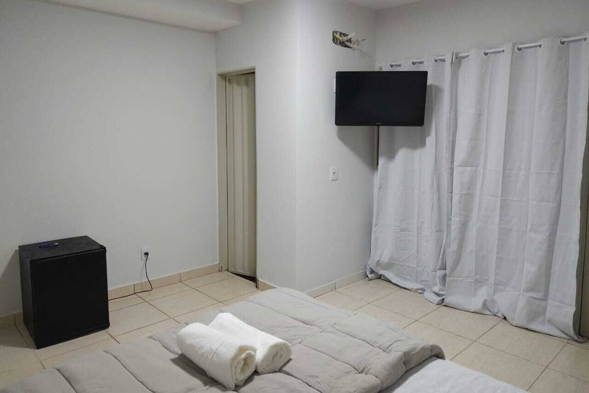 A room with a bed, a flat-screen TV mounted on the wall, a small black speaker, and a curtain.