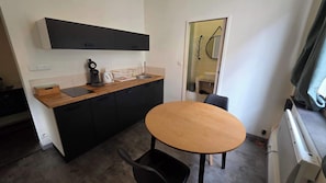 Een kleine keuken met een koffiezetapparaat, waterkoker en wastafel, een eettafeltje met stoelen, en een raam met gordijn.