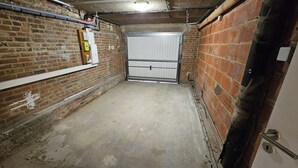 Un couloir étroit aux murs de briques et au sol en béton, avec une alarme incendie sur le côté gauche et une porte métallique fermée au bout.