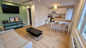 Un salon moderne avec un canapé, une table basse et un téléviseur fixé au mur. Il y a également un coin cuisine avec un tabouret de bar et un réfrigérateur.