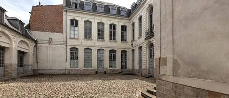 A courtyard with cobblestone flooring, a building with arched windows, and a brick roof.