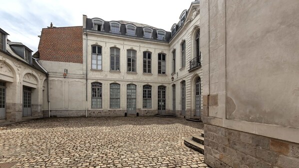 A courtyard with cobblestone flooring, a building with arched windows, and a brick roof.