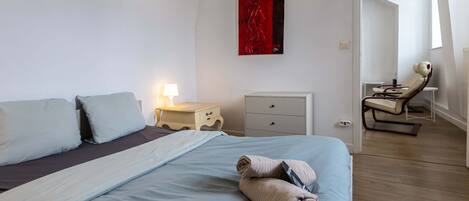 A bedroom with a bed, bedside table with lamp, a small cabinet, and a chair with a massage table.