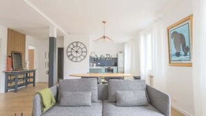 Ein modernes Wohnzimmer mit grauem Sofa, Holzboden, einem Essbereich mit rundem Tisch und einer Küche im Hintergrund.