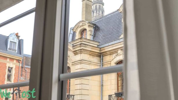 Blick durch ein Fenster eines Gebäudes im klassischen Stil, mit einem markanten Turm und aufwendigen Steinmetzarbeiten.