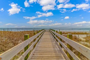 A wooden boardwalk leading to a beach with a clear sky and ocean in the background.