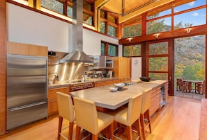 A modern kitchen with a stainless steel refrigerator, a large island with bar stools, wooden cabinetry, and large windows offering a view of a forested area. - Above It All | Luxury Mountain Estate | Hot Tub, Fire Pits, Gym + Canyon Views (Telluride)