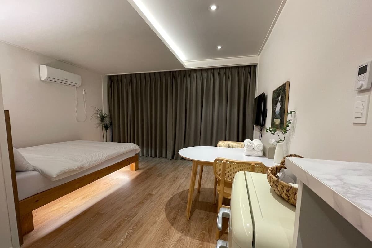 Een moderne hotelkamer met een enkel bed, een eettafel met stoelen, een kleine kitchenette en een groot raam met gordijnen.