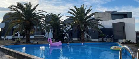 A swimming pool with a pink flamingo float, palm trees, and modern buildings in the background.