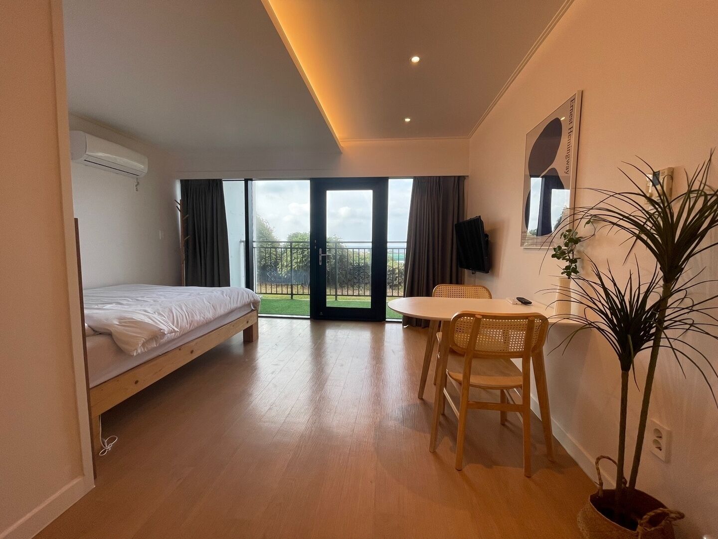 A hotel room with a bed, a desk, a chair, a potted plant, and a balcony with a view.