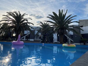 A swimming pool with inflatable toys, palm trees, and modern buildings in the background.