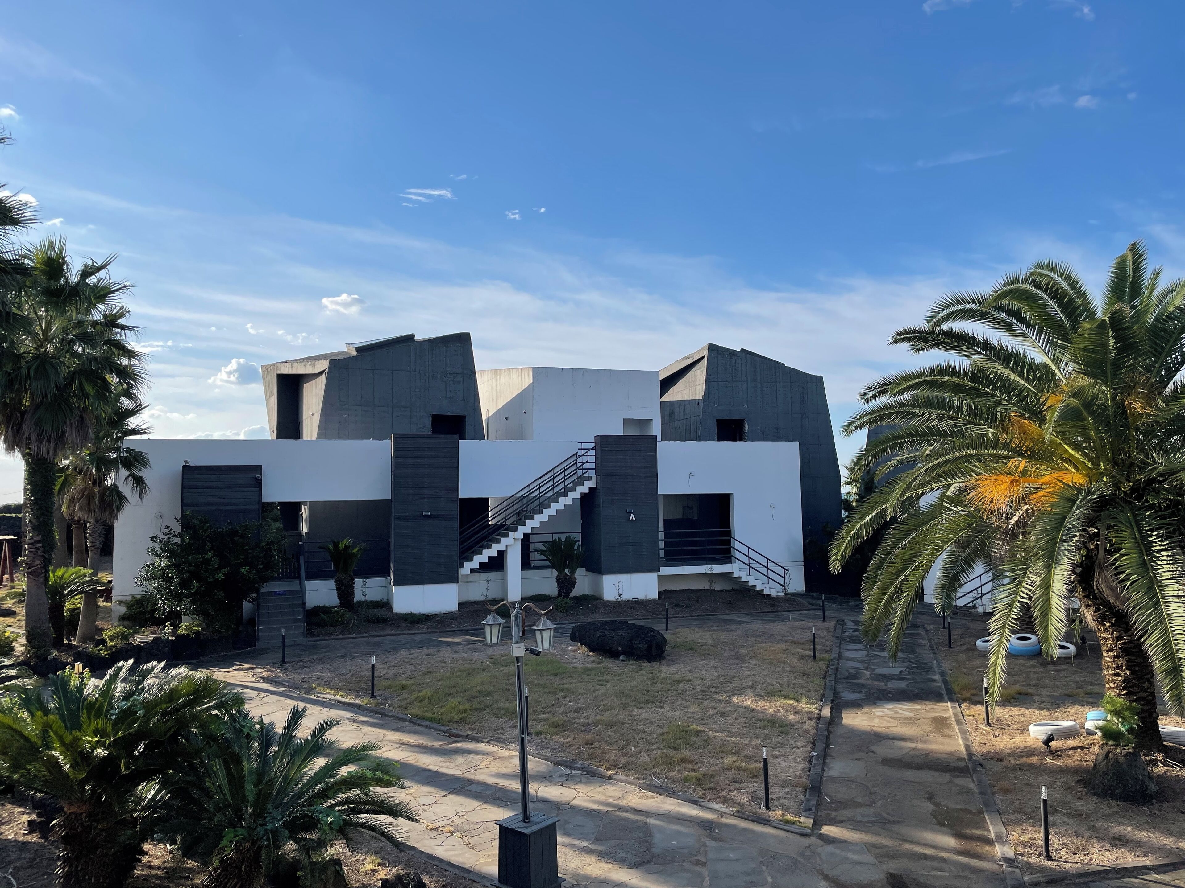 A modern building with a staircase leading to an entrance, surrounded by palm trees and a paved area.
